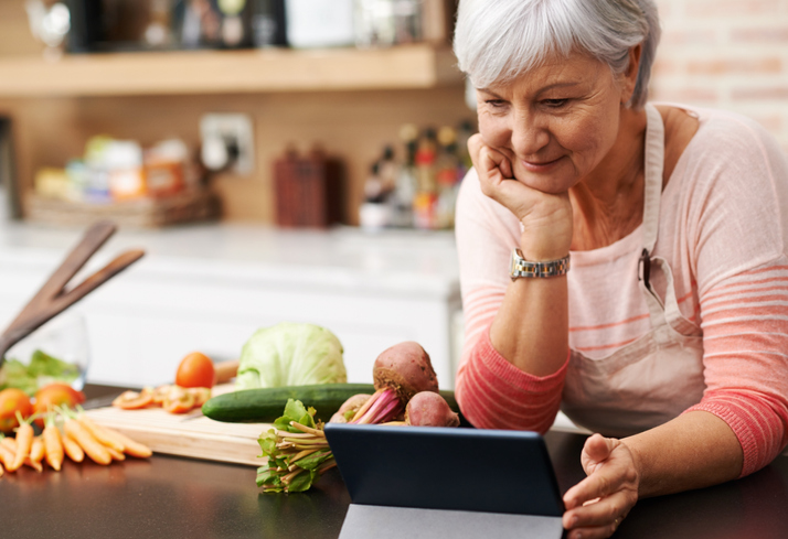 woman resting her chin on her hands as she watches a tablet in her kitchen with an assortment of foods next to her